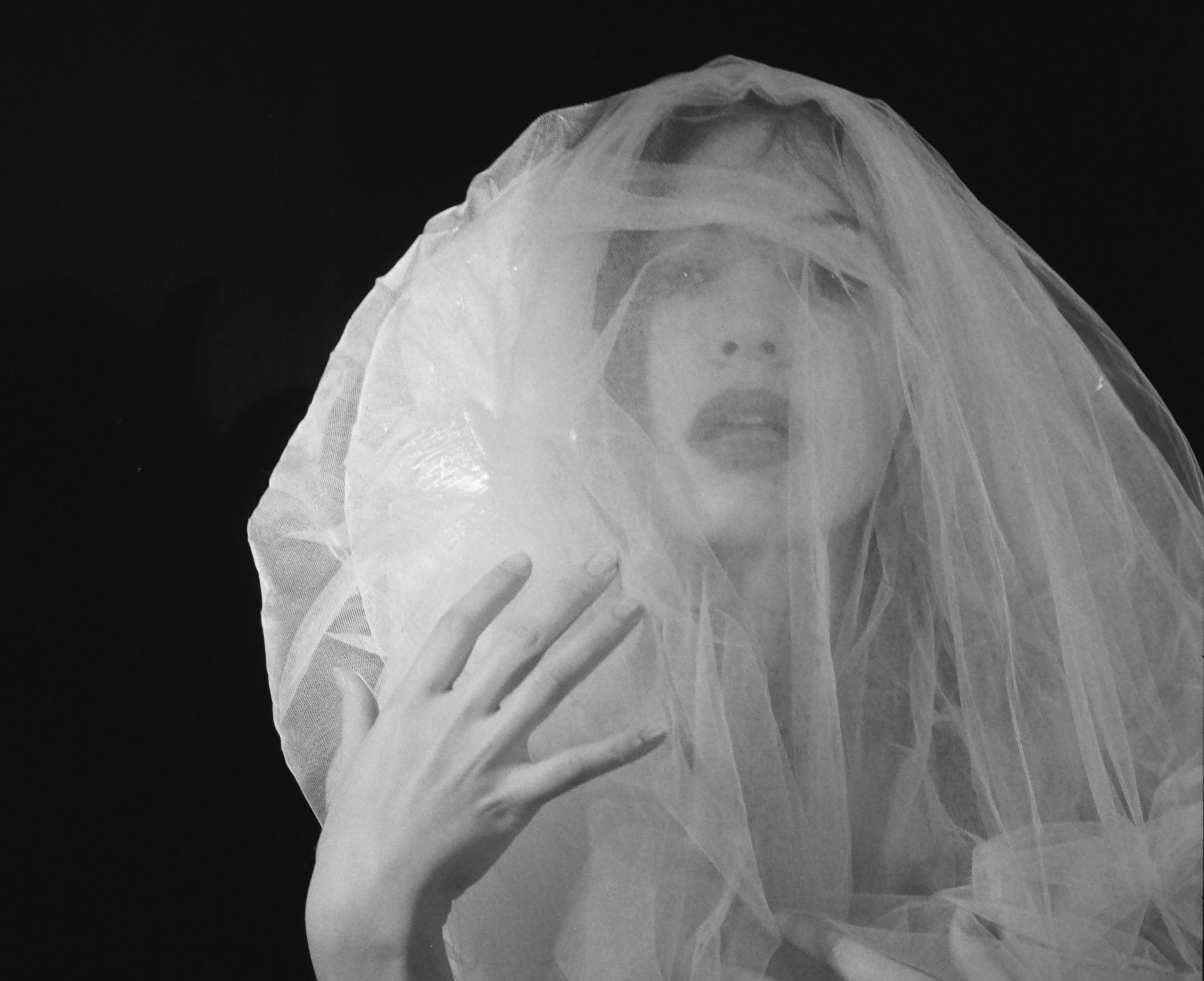Black and white close-up of a person wearing a sheer veil over the face and hand, set against a solid black background.